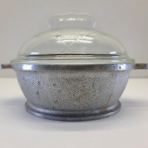 May include: A vintage, silver-colored casserole dish with a clear glass lid. The dish has a textured surface and two small handles. The lid has a decorative design. A classic kitchenware item.