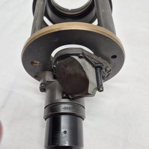 May include: A cylindrical black and dark gray metal optical component featuring a circular brass-toned mounting plate. The device includes a central lens housing, a small rectangular prism assembly, and a black barrel marked with the text 415605-40.