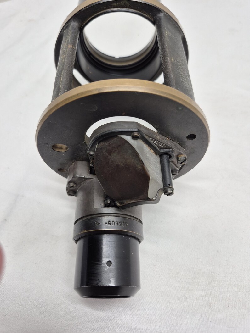 May include: A cylindrical black and dark gray metal optical component featuring a circular brass-toned mounting plate. The device includes a central lens housing, a small rectangular prism assembly, and a black barrel marked with the text 415605-40.