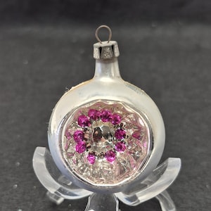 May include: Silver Christmas ornament with a circular design of pink and silver accents. The ornament has a small metal loop at the top for hanging. The ornament is displayed on a clear plastic stand.