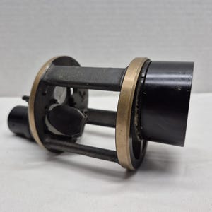 May include: A cylindrical mechanical component featuring a black metal housing and a central cage structure. The frame consists of dark gray vertical supports connected by two circular brass-colored rings, with internal black components visible inside.