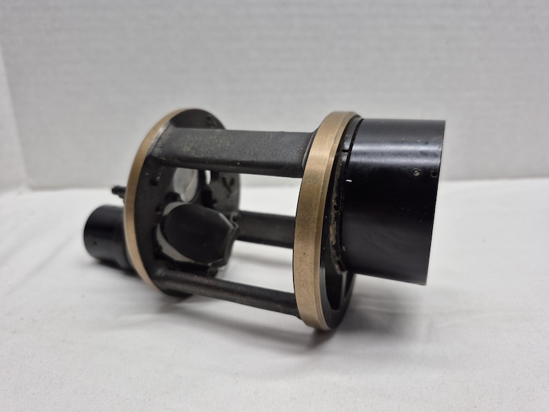 May include: A cylindrical mechanical component featuring a black metal housing and a central cage structure. The frame consists of dark gray vertical supports connected by two circular brass-colored rings, with internal black components visible inside.