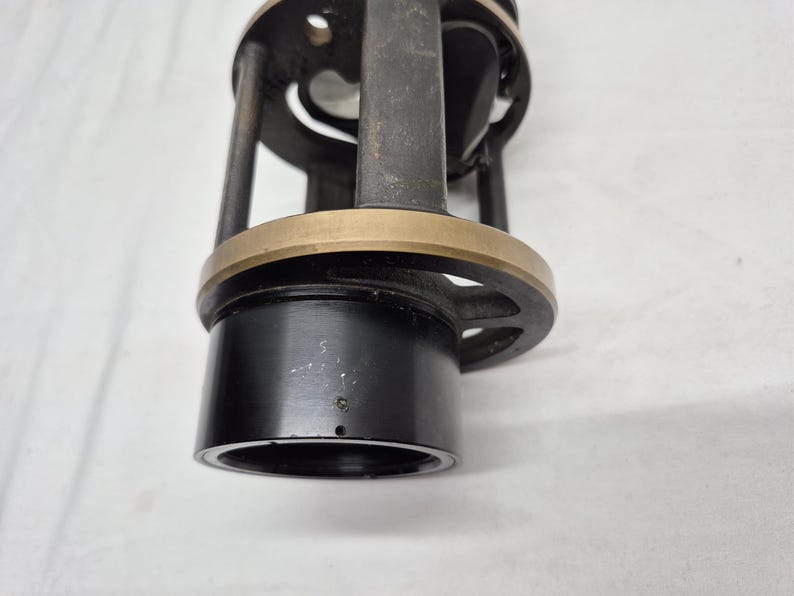 May include: A cylindrical black metal fishing reel component featuring a brass-colored circular ring, vertical support struts, and an open frame design. The base consists of a smooth black tube, while the upper section includes a circular plate.