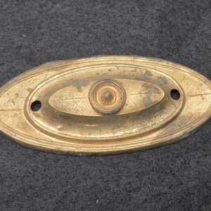 May include: An oval-shaped brass escutcheon plate featuring a central raised circular knob and a textured cross-hatched pattern. The metal hardware includes two circular screw holes on the left and right sides, with visible signs of aged patina.