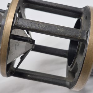 May include: A mechanical device featuring a cylindrical black housing with a brass-colored metal frame. The structure includes vertical supports and a central triangular component with the handwritten number 2 visible on its surface.