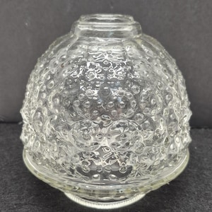 May include: A clear glass lampshade with a textured, bubble-like pattern. The shade has a rounded, bell-like shape with a slightly flared base. The glass is transparent, allowing light to pass through. The lampshade is designed to fit over a light bulb.
