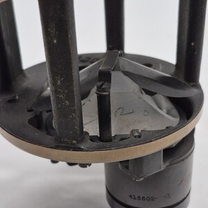 May include: A mechanical component featuring a dark gray circular frame with vertical supports and a central metallic cone. The base includes a cylindrical section with the engraved text 415502-3 and a small handwritten number 12 on the cone surface.