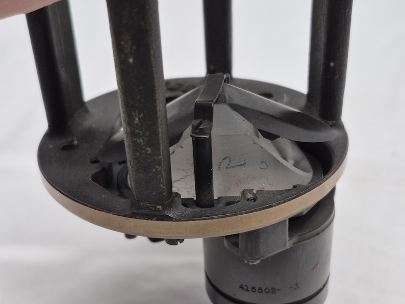 May include: A mechanical component featuring a dark gray circular frame with vertical supports and a central metallic cone. The base includes a cylindrical section with the engraved text 415502-3 and a small handwritten number 12 on the cone surface.