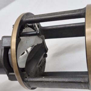 May include: A mechanical device featuring a cylindrical black housing with a brass-colored circular frame. The structure includes vertical supports and an internal white component with visible markings, secured by small black screws and metal hardware.
