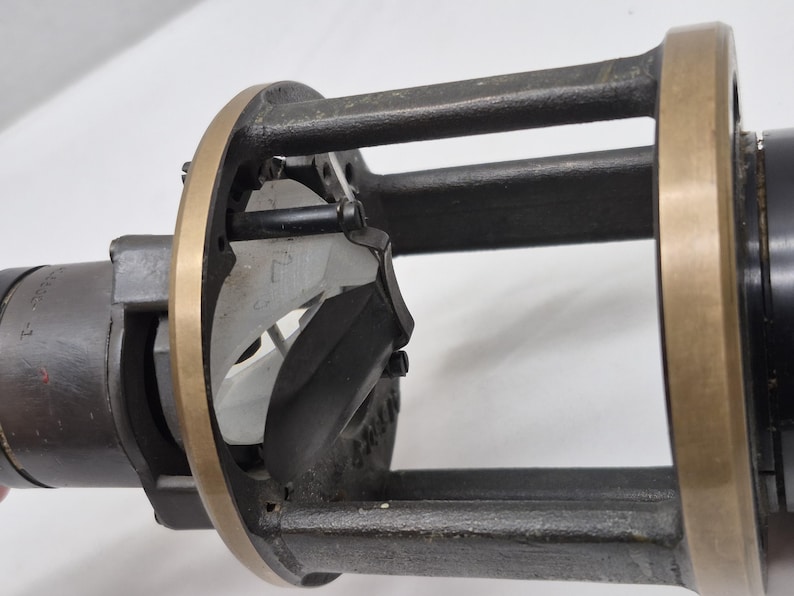 May include: A mechanical device featuring a cylindrical black housing with a brass-colored circular frame. The structure includes vertical supports and an internal white component with visible markings, secured by small black screws and metal hardware.