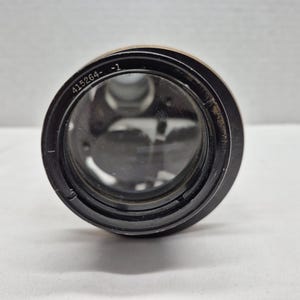 May include: A black camera lens with a circular glass element. The outer rim of the lens features the engraved text 415264- -1. The interior of the lens shows visible mechanical components and glass elements through the front glass surface.