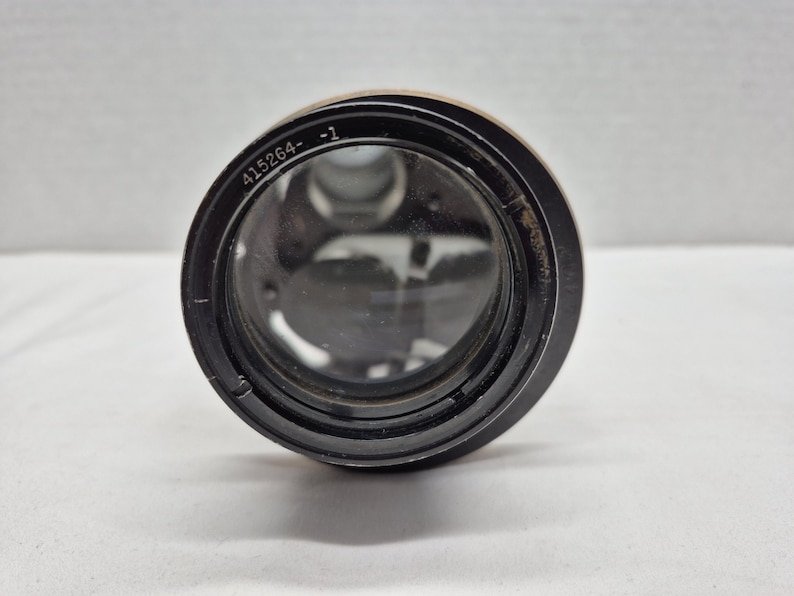 May include: A black camera lens with a circular glass element. The outer rim of the lens features the engraved text 415264- -1. The interior of the lens shows visible mechanical components and glass elements through the front glass surface.