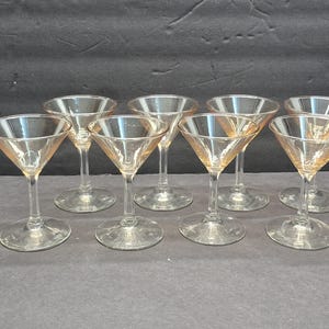 May include: Set of eight vintage martini glasses. Each glass features a classic conical bowl and a slender stem, with a round base. The glasses have a subtle amber tint, adding a touch of warmth. Perfect for cocktails and entertaining.