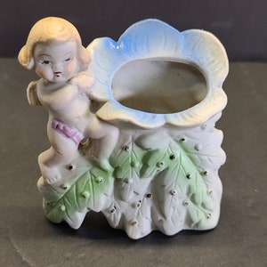 May include: A white ceramic planter shaped like a flower with a cherub climbing on the side. The flower has green leaves and blue petals. The cherub is wearing a pink sash.