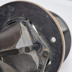 May include: A mechanical component featuring a circular black metal frame with a metallic rim. A central glass prism is secured by a black bracket held with screws. The bracket is engraved with the serial number 415251. A small circular hole is visible on the right side of the frame.