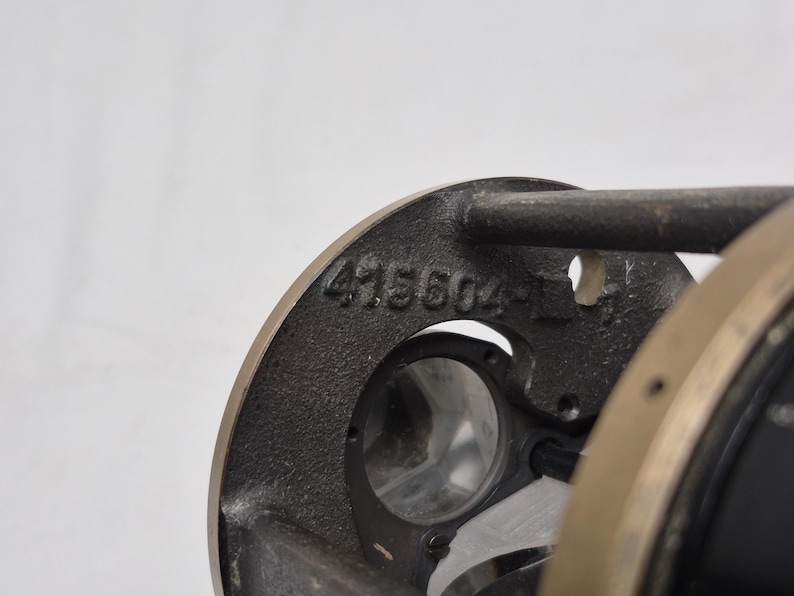 May include: A dark gray cast metal component featuring a circular frame with the embossed number 415604-1. The object includes a central opening with visible internal glass elements and a smooth, metallic outer edge, showing industrial construction.