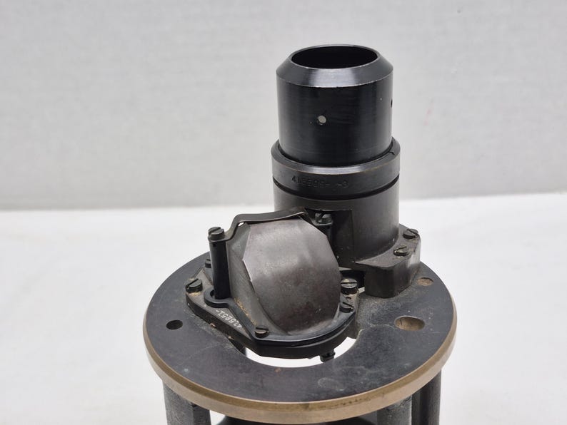 May include: A black cylindrical optical component featuring a vertical tube and a curved, metallic reflective prism assembly mounted on a circular base. The assembly includes small screws and engraved numerical markings on the metal surfaces.