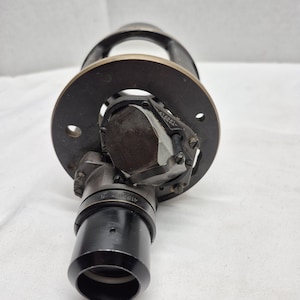 May include: A black metal optical instrument component featuring a circular mounting plate with two holes, a central glass prism assembly, and a cylindrical lens barrel. The metal frame displays the engraved serial number 415251 on the prism housing.