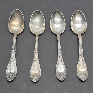 May include: A set of four silver-toned metal spoons arranged in a row. Each spoon features an oval bowl and a handle decorated with intricate embossed floral and leaf patterns, tapering toward a wider, rounded base with detailed engravings.