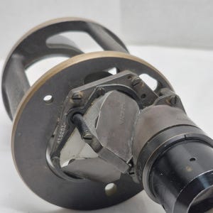 May include: A vintage optical instrument featuring a circular dark metal mounting plate with a brass-toned rim. A central glass prism assembly is secured by a metal frame marked with the number 415251. A black cylindrical lens housing extends outward.