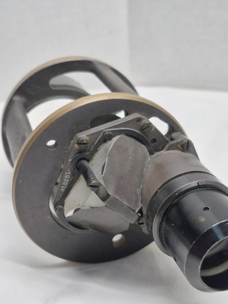May include: A vintage optical instrument featuring a circular dark metal mounting plate with a brass-toned rim. A central glass prism assembly is secured by a metal frame marked with the number 415251. A black cylindrical lens housing extends outward.