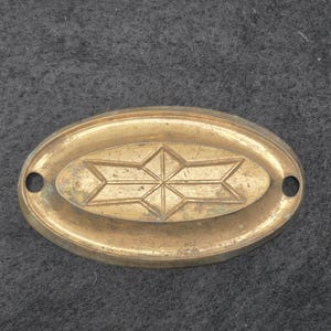 May include: An oval-shaped brass plate featuring a central embossed geometric star pattern. The plate has a smooth, metallic finish with visible signs of aging and patina. It includes two circular mounting holes, one on each side of the oval.
