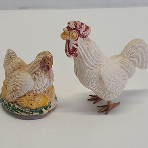 May include: Two white ceramic figurines of a rooster and a hen. The rooster has a red comb and wattle and a white body with a brown tail. The hen is sitting on a nest of yellow eggs.