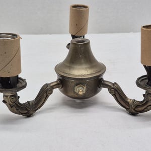 May include: A brass-toned metal light fixture base featuring three upward-facing bulb sockets, each covered with a light brown cardboard sleeve. The central hub has a rounded dome shape with a small metal switch, and two curved arms extend outward.