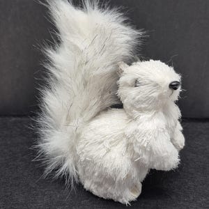 May include: A white, plush squirrel figurine with a fluffy tail and body. The squirrel has a black nose and small, closed eyes. The fur has a slight texture and some strands of gray. The figurine is set against a dark background.