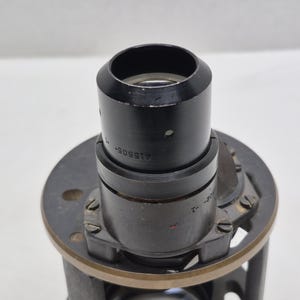 May include: A black cylindrical optical lens component featuring a metallic mounting plate with small screws. The side of the lens barrel is engraved with the text 415505-1. The structure is composed of dark metal with a circular aperture at the top.