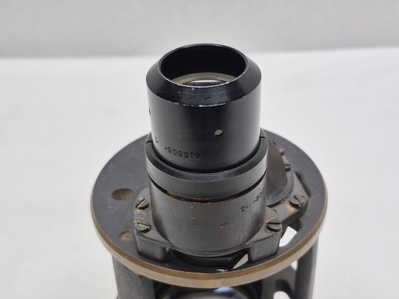 May include: A black cylindrical optical lens component featuring a metallic mounting plate with small screws. The side of the lens barrel is engraved with the text 415505-1. The structure is composed of dark metal with a circular aperture at the top.
