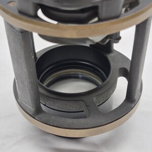 May include: A cylindrical mechanical component featuring a dark gray metal frame with vertical supports and a circular bronze-toned top ring. The interior contains a black circular housing with a central opening, showcasing a layered industrial design.