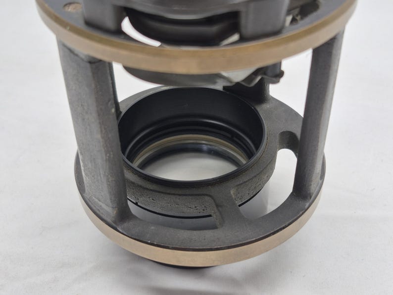 May include: A cylindrical mechanical component featuring a dark gray metal frame with vertical supports and a circular bronze-toned top ring. The interior contains a black circular housing with a central opening, showcasing a layered industrial design.