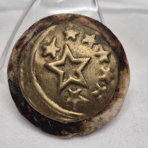 May include: A circular button featuring a brass-toned metal inlay with a crescent moon and multiple stars embossed on the surface. The outer edge of the button is composed of a textured, organic material with brown and cream-colored markings.