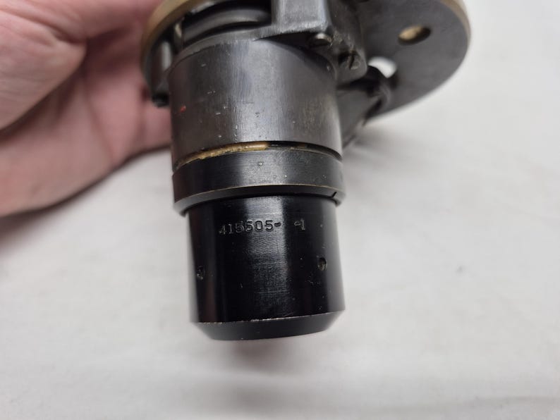 May include: A cylindrical black metal mechanical component featuring a brass-colored top flange and a series of small circular indentations. The side of the lower section is clearly stamped with the alphanumeric text 415505-1 in a clean, sans-serif font.