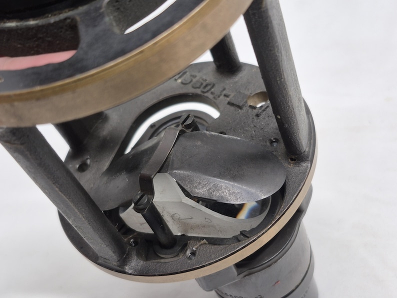 May include: A mechanical optical component featuring a dark gray metal frame with a brass-toned ring. The assembly includes a central prism with a curved metal shutter mechanism and a small screw. The number 1550-J-1 is engraved on the frame.