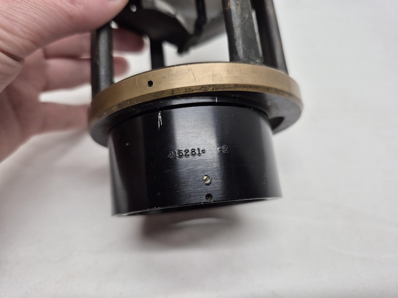 May include: A cylindrical black metal component featuring a brass-colored ring at the top and vertical support struts. The black base displays the engraved text 415261-2, along with small circular holes and a tiny metal pin protruding from the surface.
