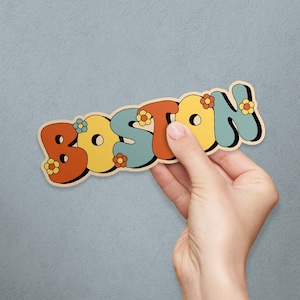 May include: A colorful sticker with the word "Boston" in a retro font with yellow flowers. The sticker is on a gray wall.