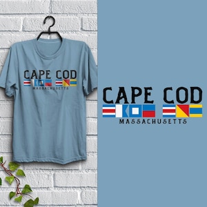 May include: Light blue t-shirt with the words "CAPE COD MASSACHUSETTS" in black. The text is above a row of colorful nautical signal flags. The shirt is hanging on a black hanger against a white brick wall.