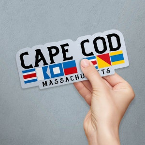 May include: A sticker with the words "CAPE COD MASSACHUSETTS" in black letters. The sticker has a light gray background and features nautical signal flags. The sticker is held by a hand against a gray background.