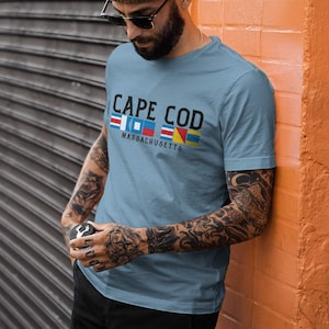 May include: A light blue t-shirt with the words "CAPE COD MASSACHUSETTS" in black. The text is above a row of colorful nautical flags. The shirt is short-sleeved.