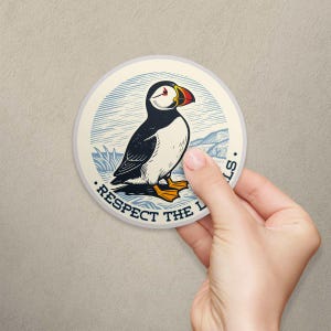 May include: A circular sticker featuring a black, white, and orange puffin illustration set against a blue and cream landscape background. The text "RESPECT THE PUFFINS" is printed in a dark blue font along the bottom edge of the circular design.