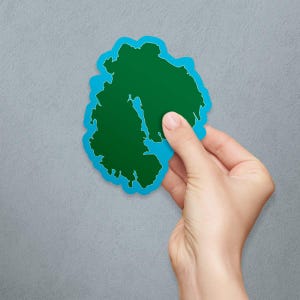 May include: A green island sticker with a blue outline. The sticker is being held by a hand against a gray background.