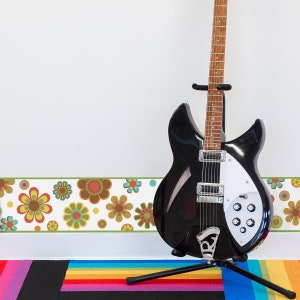 May include: A black electric guitar with a white pickguard and a floral patterned border along the bottom of the wall. The guitar is on a black stand with a rainbow rug underneath.