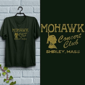 May include: Dark green t-shirt with the text "MOHAWK Concert Club SHIRLEY, MASS" in a distressed gold font. The design includes a silhouette of a Native American figure. The shirt is hanging on a black hanger against a white brick wall.