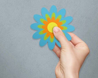 May include: A hand holding a blue paper flower with a yellow, orange, and green center.