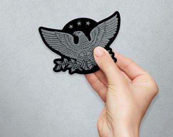 May include: A black and gray eagle emblem with outstretched wings, three stars, and a laurel branch. The eagle is a detailed illustration, held in a hand against a neutral background.
