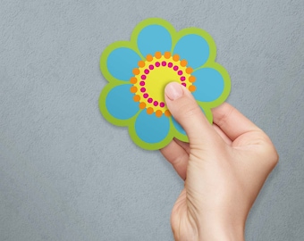May include: A colorful flower-shaped sticker with a blue and green design, held by a hand. The flower has a yellow center with pink and orange dots. The sticker is against a gray background.