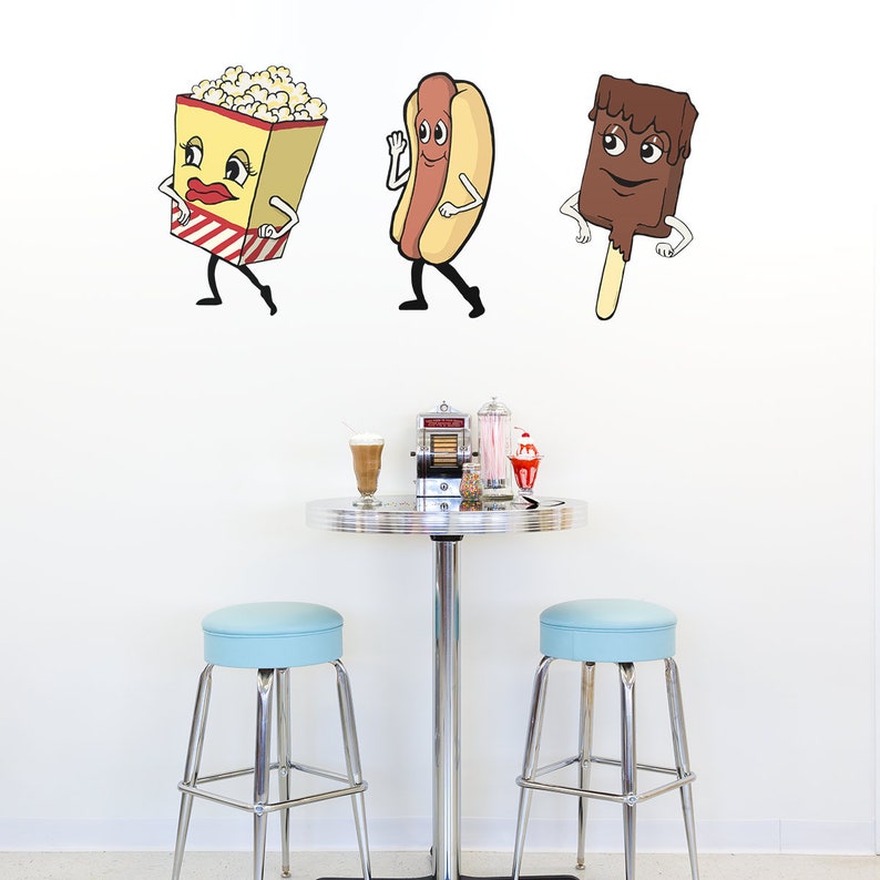 May include: Three cartoon food characters, a popcorn bucket, a hot dog, and an ice cream bar, are on a white wall. The characters are all smiling and have arms and legs. There are two blue stools and a silver table with a silver milkshake mixer, a glass of milkshake, and a red and white striped cup with a red and white straw on the table.