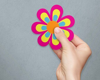 May include: A pink flower with yellow and blue petals and a bright orange center. The flower is being held by a hand against a gray background.